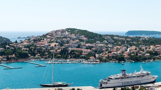 Un pueblo costero con un puerto deportivo, un gran crucero llamado 'Jadrolinja', y un paisaje montañoso de fondo.