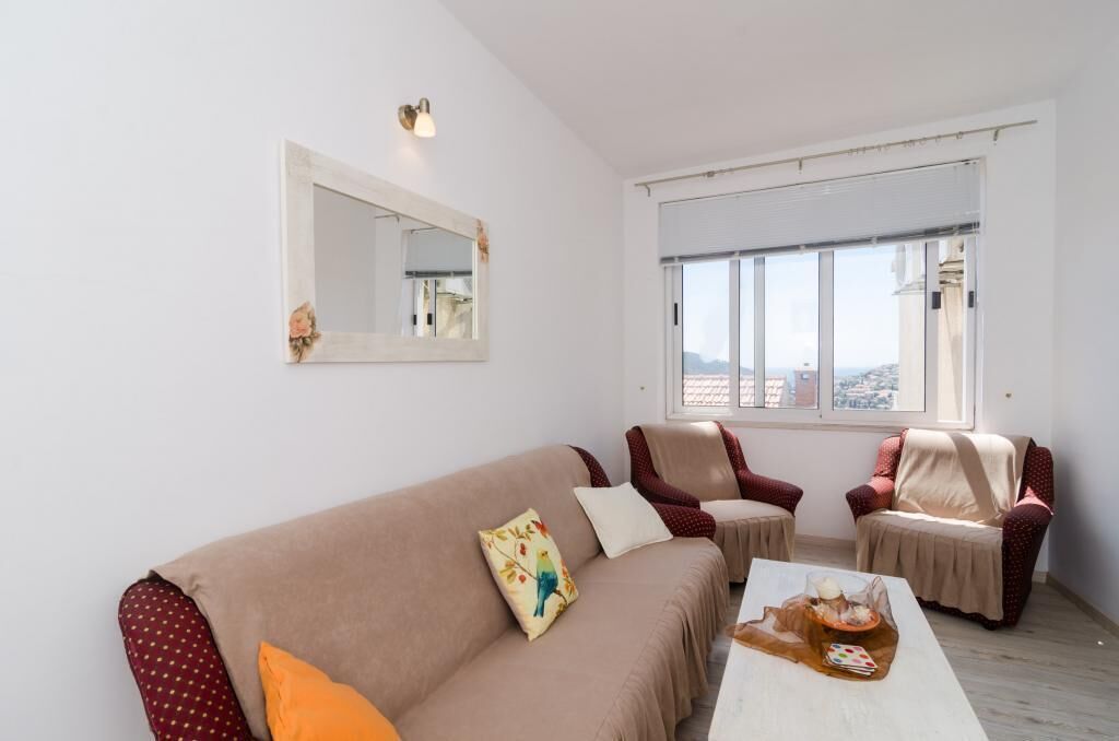 A living room with a beige sofa, two armchairs, a coffee table, a large window with blinds, and a wall-mounted mirror.