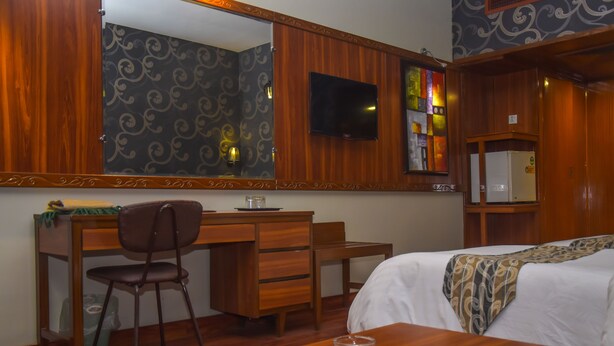 A hotel room with a wooden desk, a chair, a bed with a patterned blanket, a TV mounted on the wall, and a refrigerator. - Hotel Faran (Karachi)