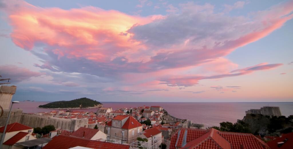 Eine Küstenstadt mit roten Dächern, einer markanten Festung und Blick auf das Meer unter einem rosa-blauen Himmel.