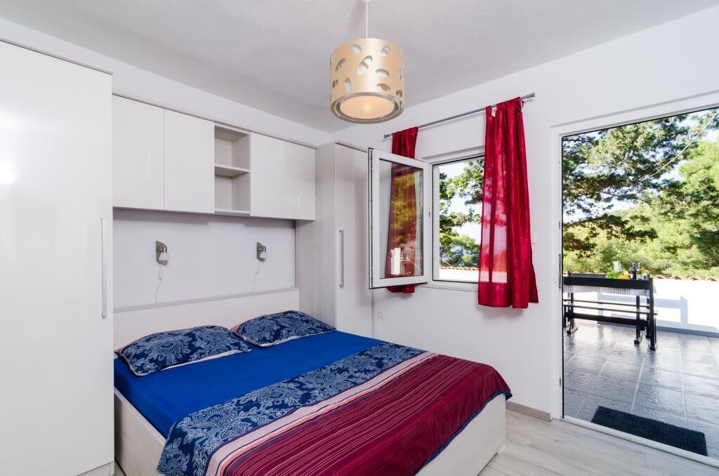 Ein Schlafzimmer mit einem Bett, einem Fenster mit roten Vorhängen und Blick auf Grünflächen.