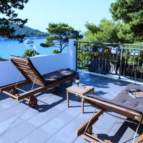 A balcony with a wooden lounge chair and a small table, overlooking a body of water and trees.