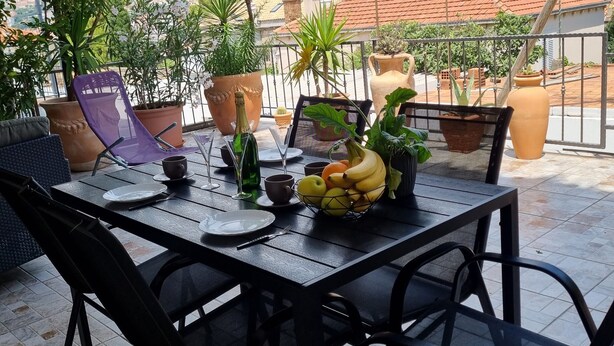 A terrace with a table set for two, surrounded by potted plants and a view of a hillside town. - Apartments Mavi - Two-Bedroom Apartment with Terrace and City View (Dubrovnik)