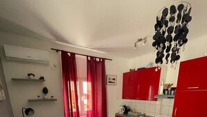 A compact living space with a dining area, a sofa, a small kitchen with red cabinets, and a ceiling light fixture. - Apartments Mavi - Two-Bedroom Apartment with Terrace and City View (Dubrovnik)