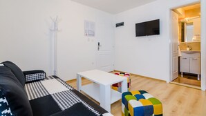 Una sala de estar con un sofá, una mesa blanca, un reposapiés colorido, un perchero y un televisor montado en la pared.