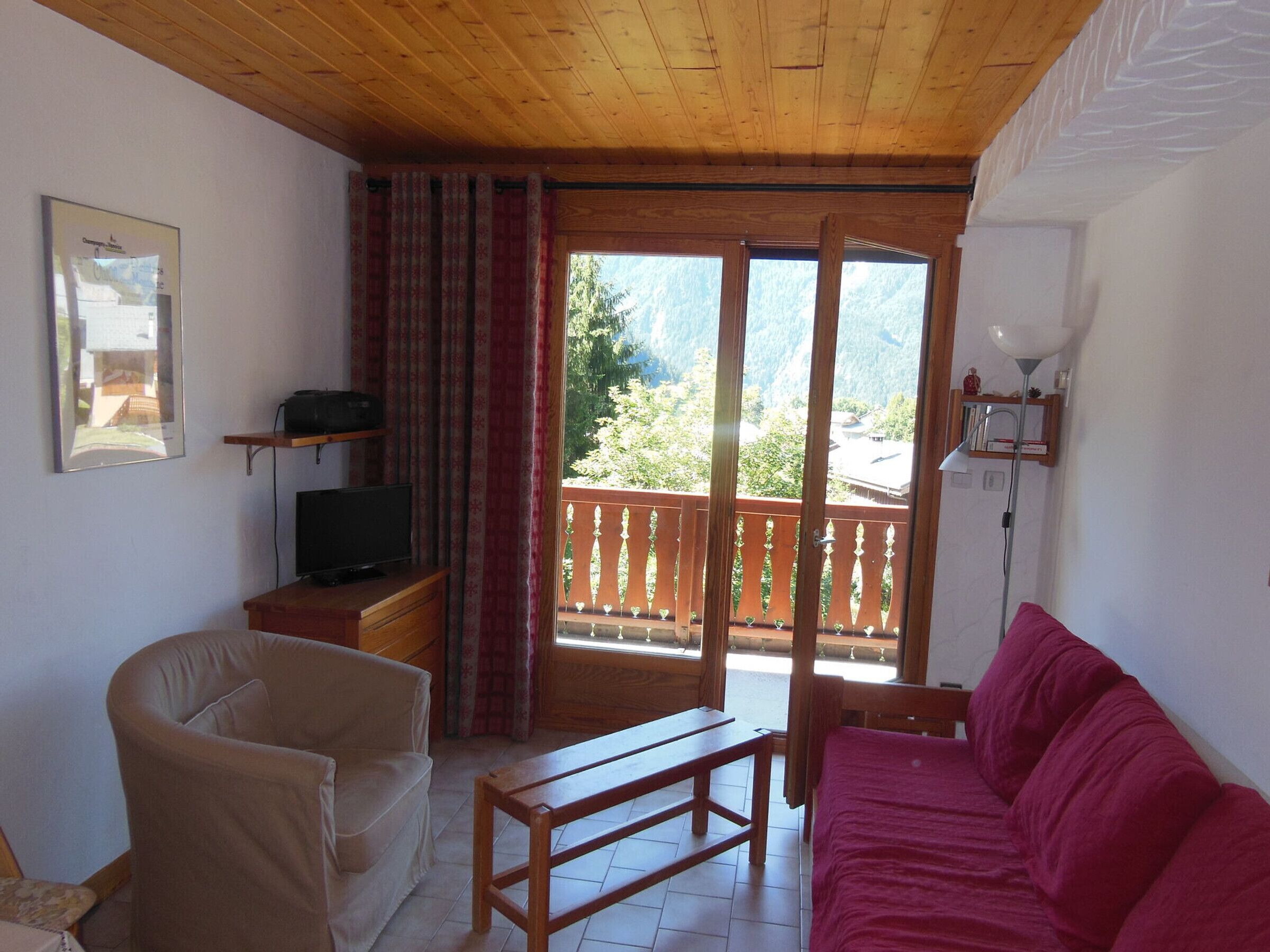 Quarto com teto de madeira, sofá vermelho, poltrona bege, mesa de madeira, televisão sobre um suporte e varanda com grades de madeira.