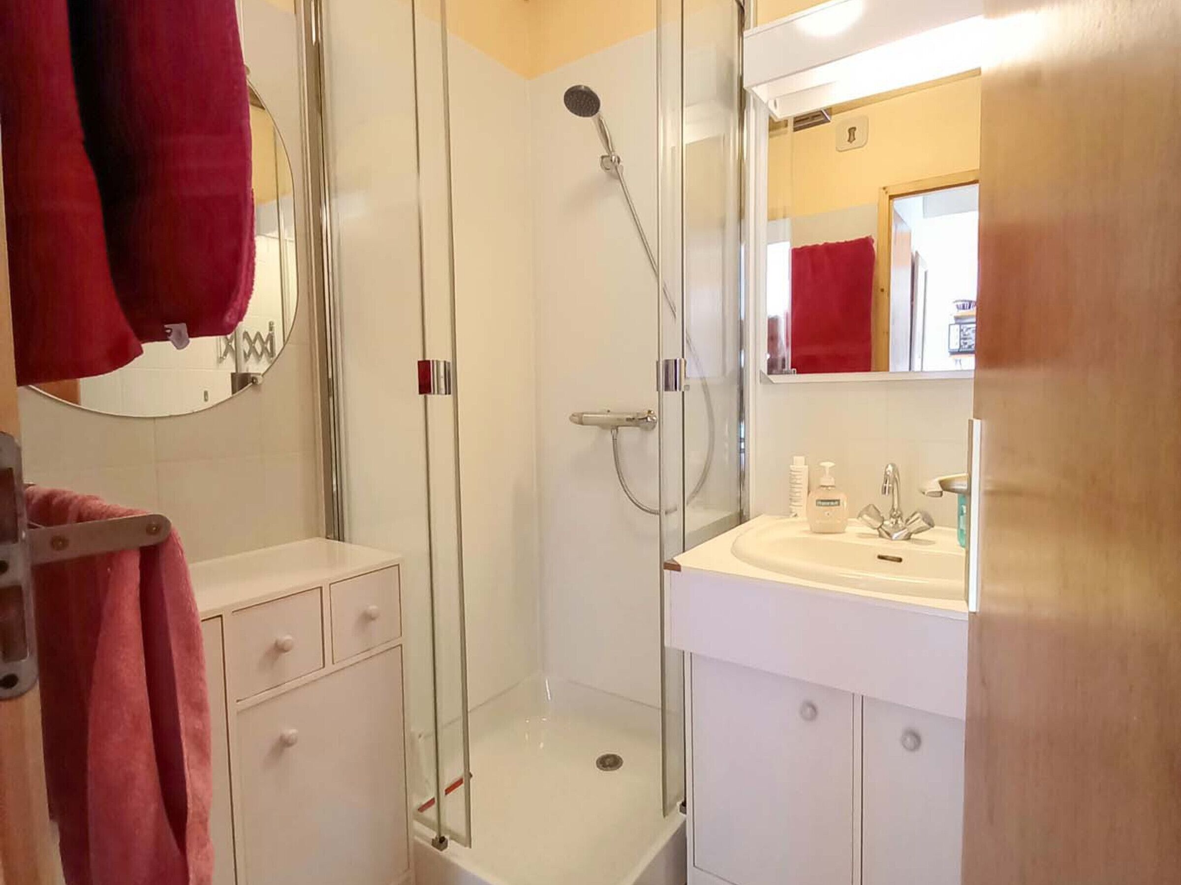 Un bagno con doccia vetrata, un lavandino bianco con uno specchio sopra e un asciugamano rosso su un portasciugamani.