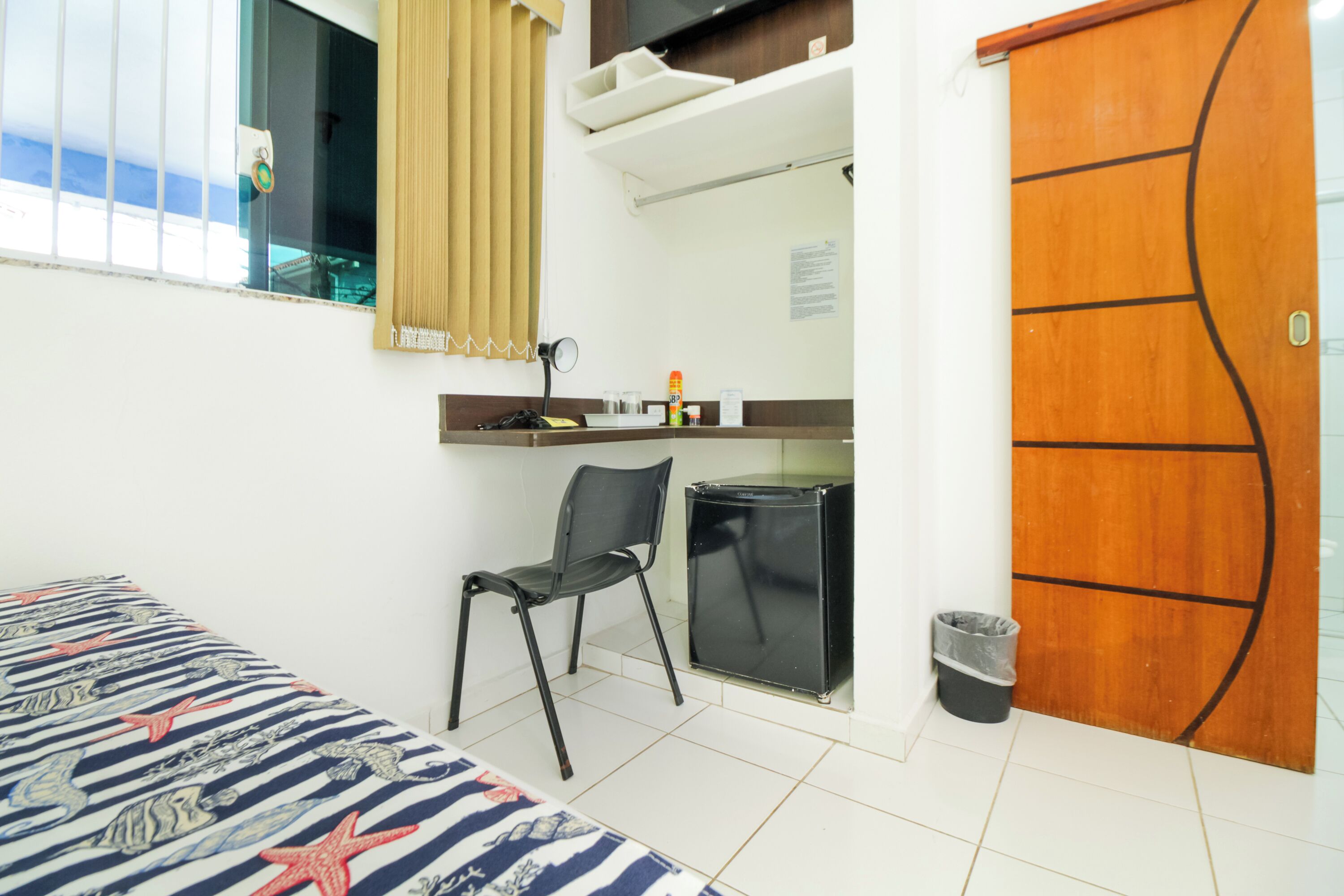 Quarto de hotel com uma cama, escrivaninha, cadeira, mini-geladeira, lixeira e porta de madeira.