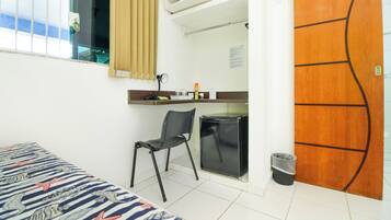 Quarto de hotel com uma cama, escrivaninha, cadeira, mini-geladeira, lixeira e porta de madeira.
