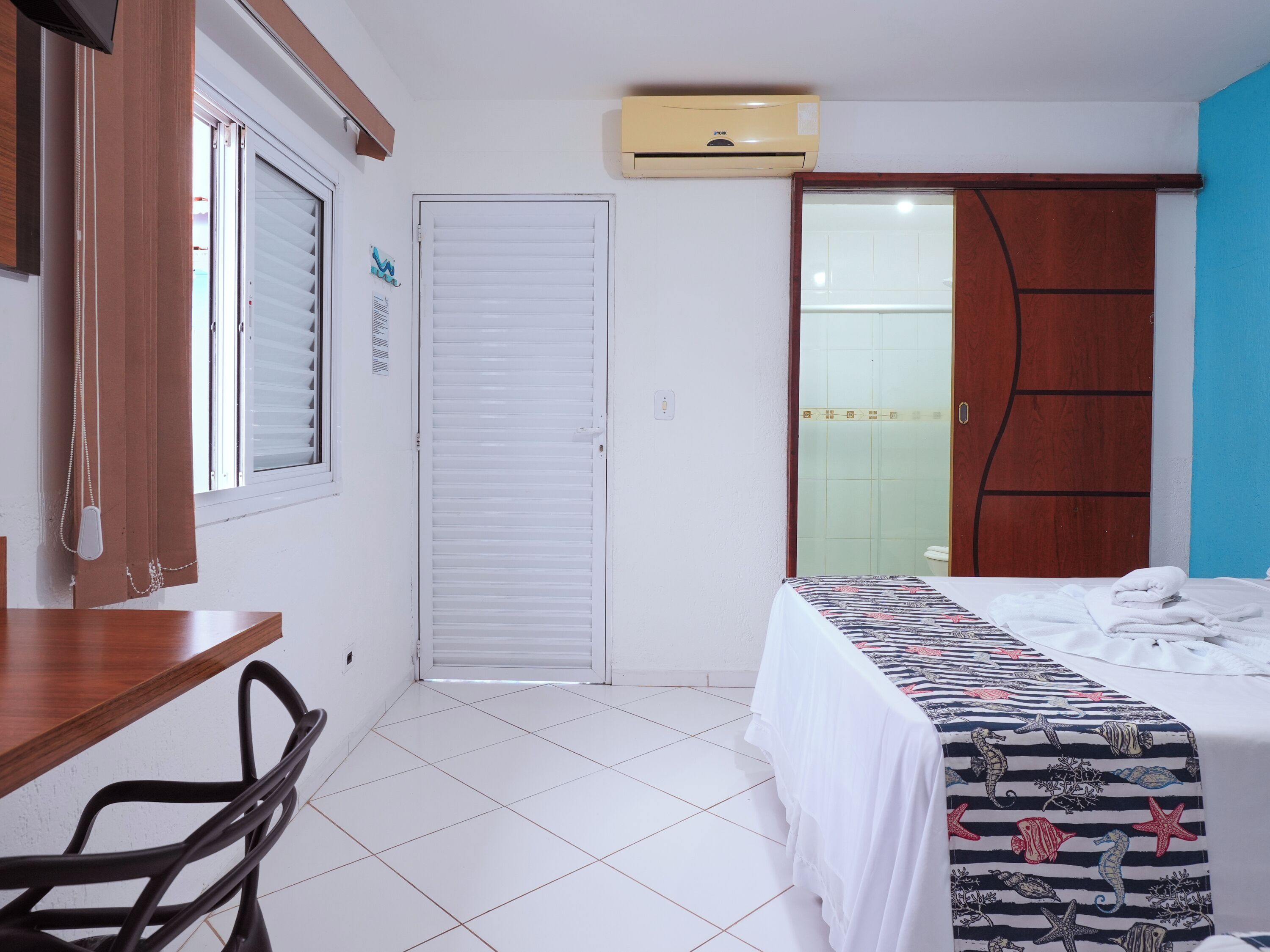 A hotel room with a bed, a desk, a chair, a wardrobe, and a bathroom with a glass door.