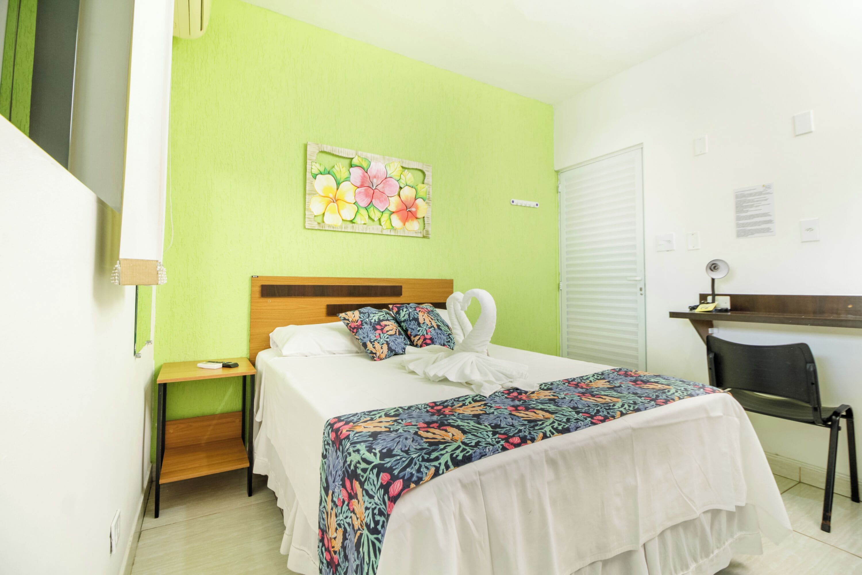 Habitación de hotel con cama, escritorio, silla, mesita de noche y pared verde con una pintura floral.