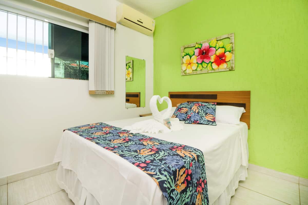 Ein Hotelzimmer mit einem Bett, einem Fenster mit Jalousien, einer Wandklimaanlage und einem Blumenbild.