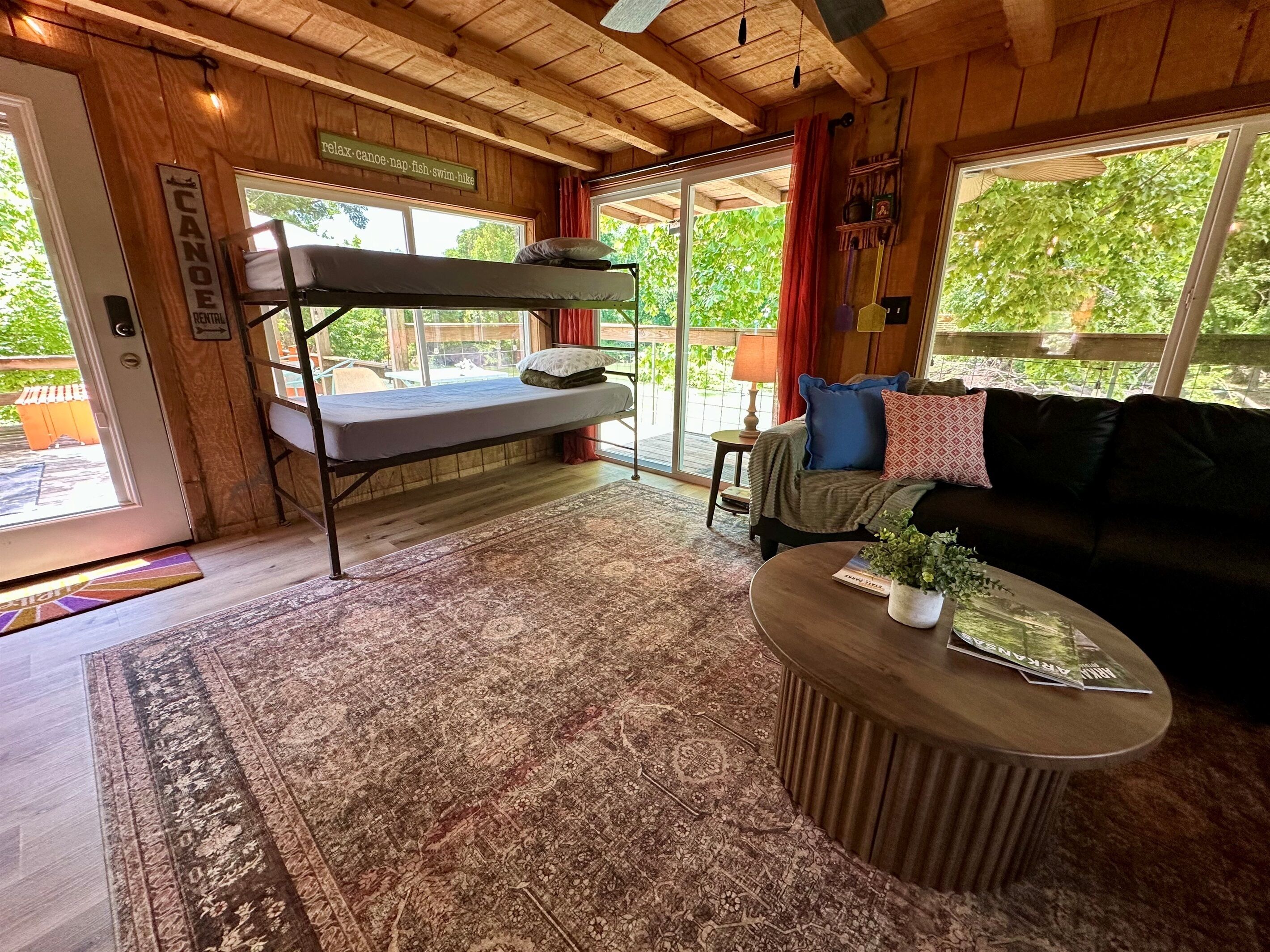 A cozy cabin interior with a bunk bed, a sofa, a coffee table, and a large window offering a view of greenery.