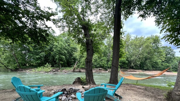 Een camping met turquoise Adirondack stoelen en een hangmat nabij een rivier, omgeven door bomen.