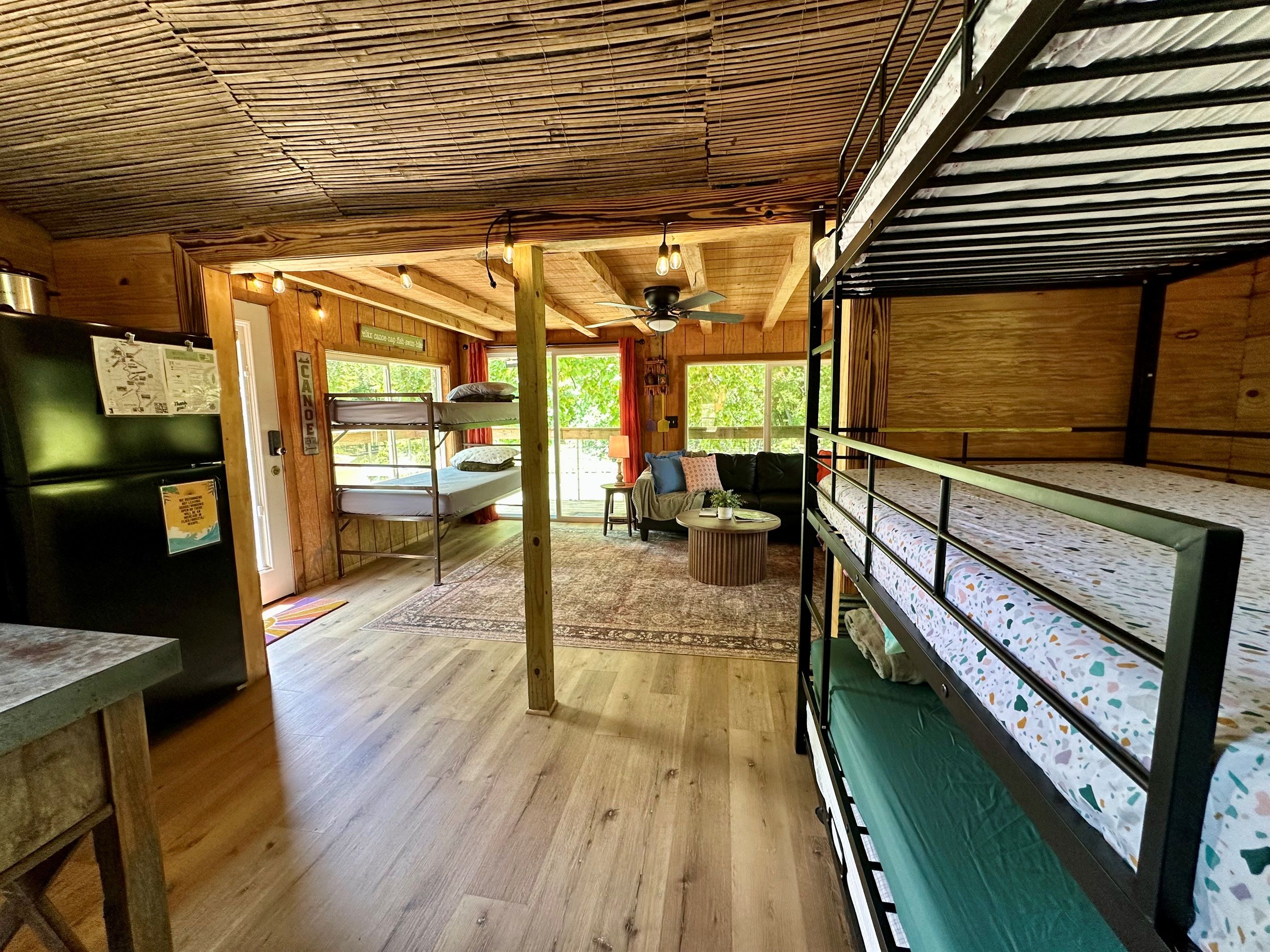 A rustic cabin interior with wooden walls, a bunk bed, a dining table, and a ceiling fan.