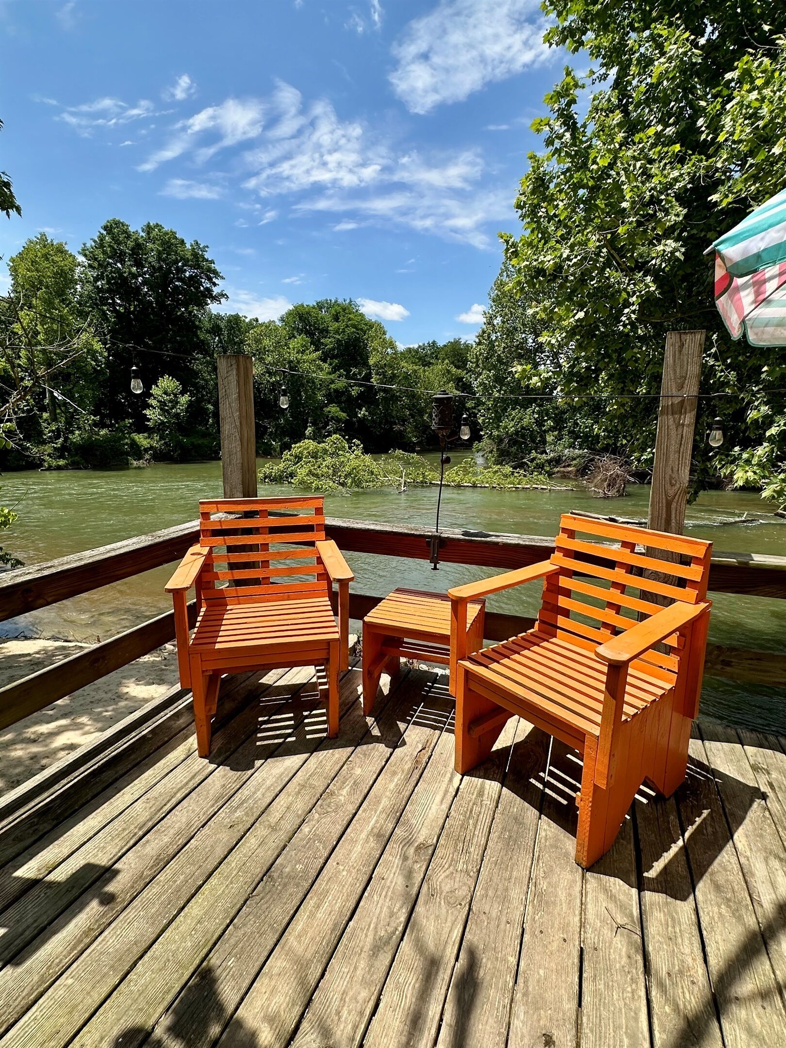 Une terrasse en bois avec deux chaises en bois et une petite table, donnant sur une rivière bordée d’arbres.