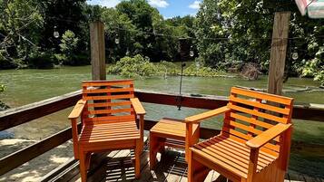 Une terrasse en bois avec deux chaises en bois et une petite table, donnant sur une rivière bordée d’arbres.