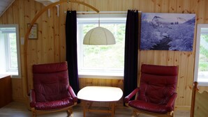 A room with wooden walls, a window with curtains, a painting, and two red chairs around a small table. - Lunheim Accomodation (Stranda)