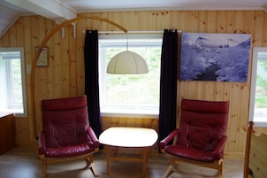 Una habitación con paredes de madera, una ventana con cortinas, un cuadro y dos sillas rojas alrededor de una mesa pequeña.