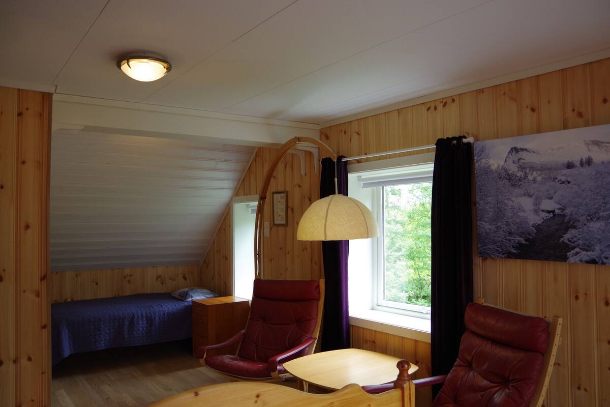 A cozy cabin room with a bed, two chairs, a table, and a large window with a view of greenery.