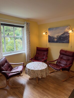 A room with a wooden floor, a round table with a floral tablecloth, two leather armchairs, a window with a view of greenery, and a painting on the wall. - Lunheim Accomodation (Stranda)