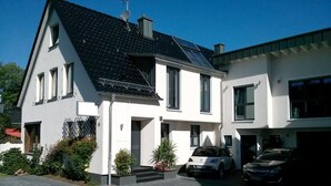 A modern two-story house with a sloped roof, solar panels, and a driveway with two cars parked.