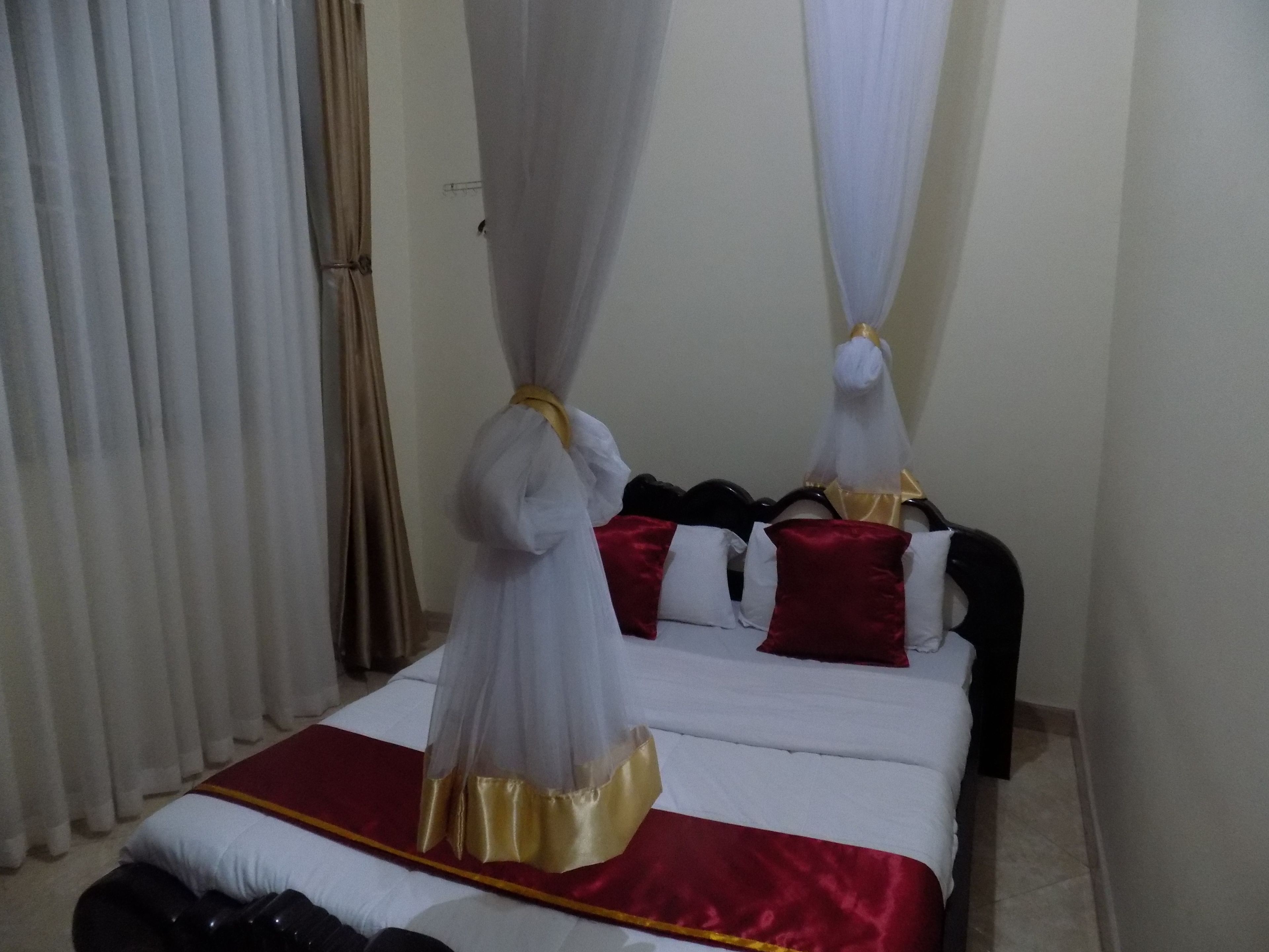 Habitación de hotel con una cama, cortinas blancas, una colcha roja y dorada, y un dosel.