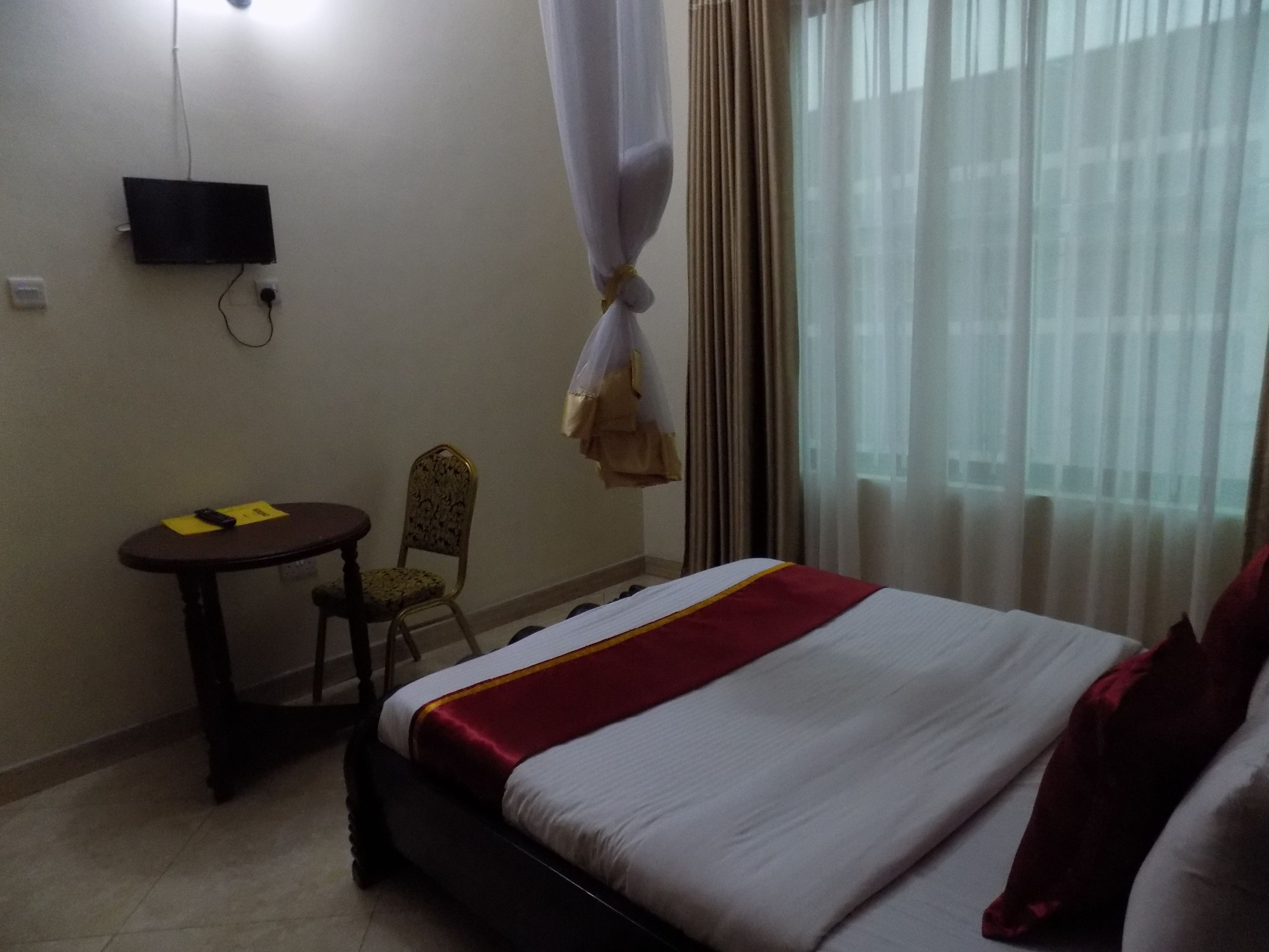 A hotel room with a bed, a small table, a chair, a TV mounted on the wall, and a window with curtains.