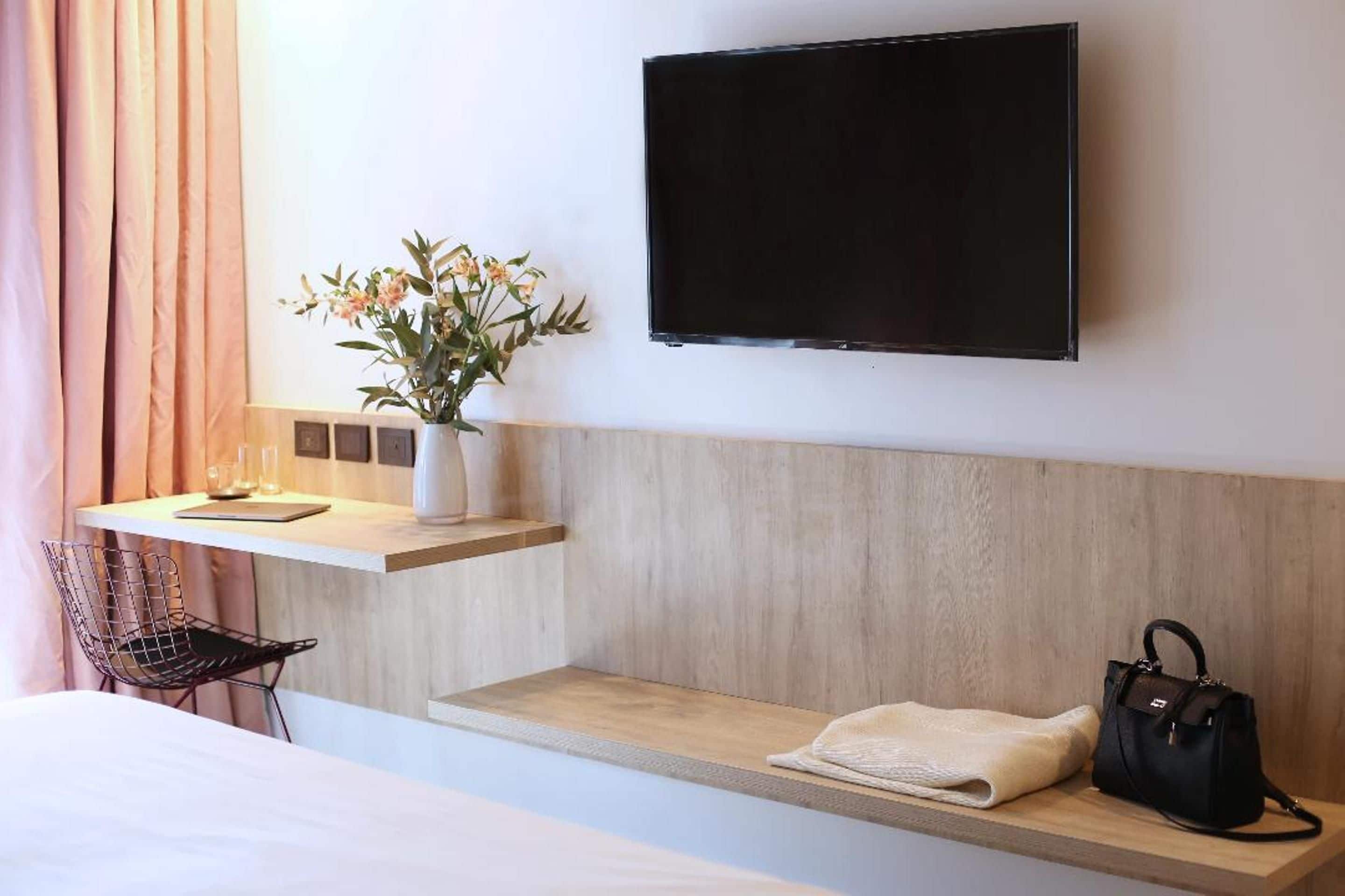 A hotel room with a bed, a desk with a chair, a TV mounted on the wall, a shelf with a vase of flowers, and a bench with a bag and towel.