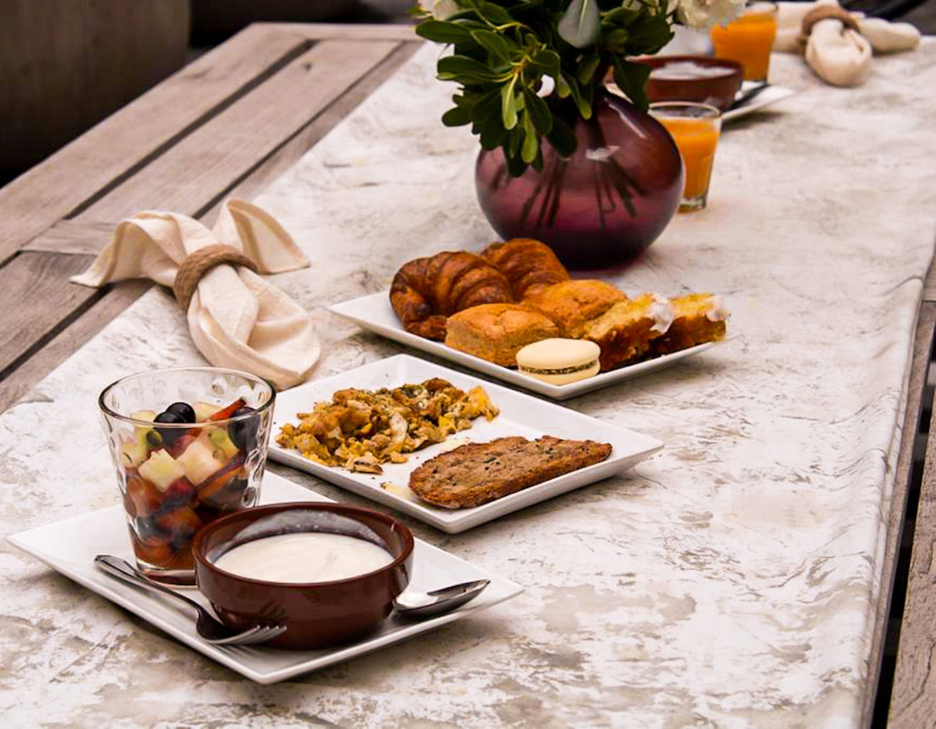 Una mesa de desayuno con cruasanes, un tazón de sopa, un vaso de jugo y una ensalada de frutas.