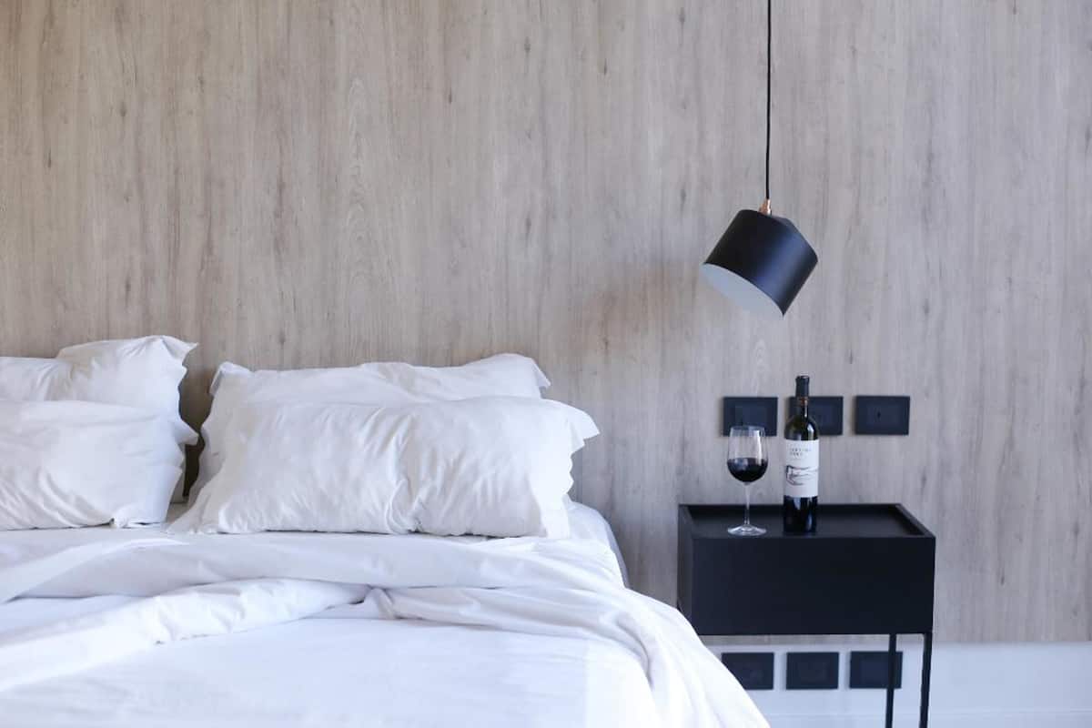 A neatly made bed with white linens, a black bedside table with a wine bottle and glass, and a modern pendant light.