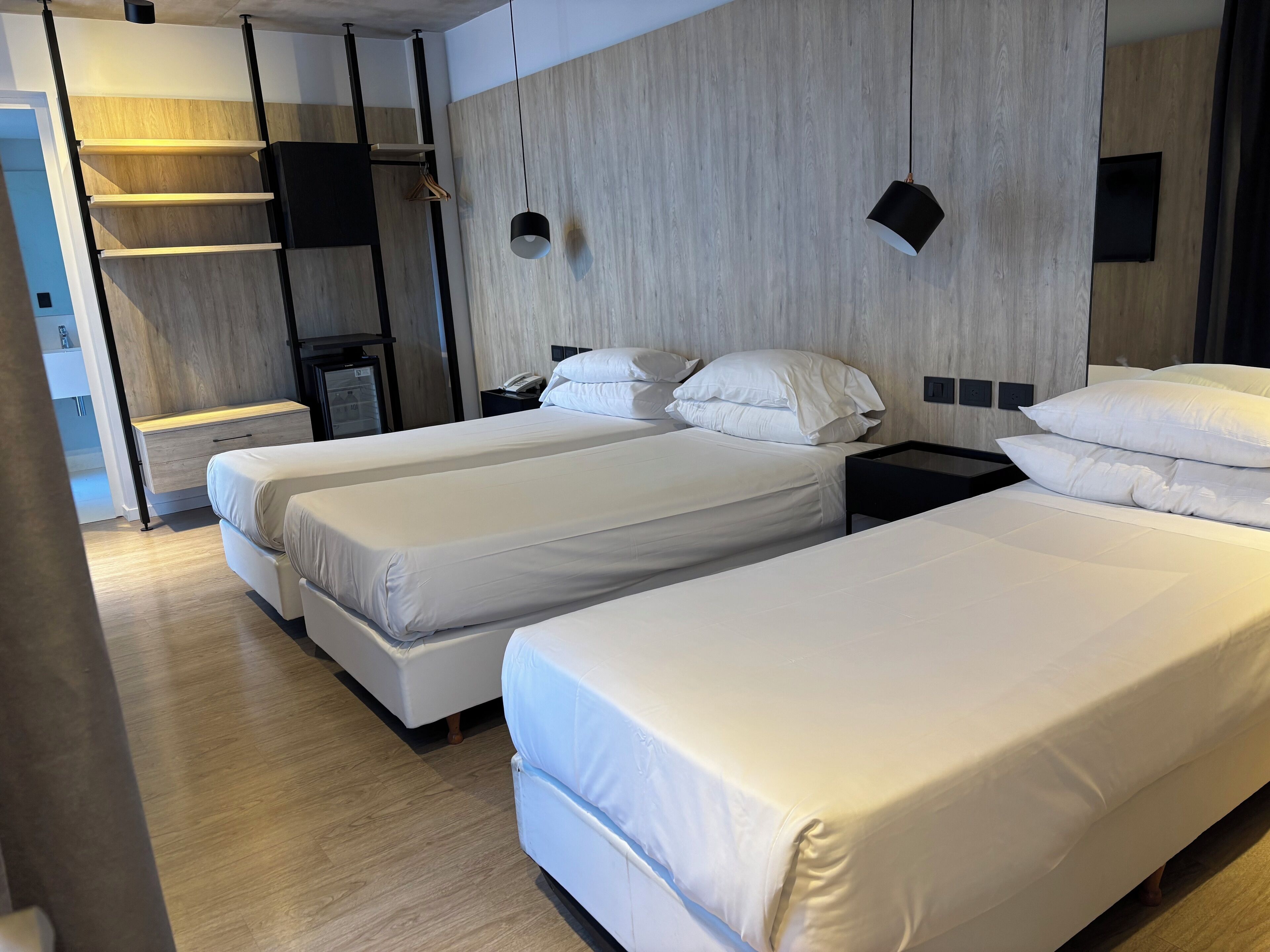 Habitación de hotel con dos camas individuales, cabecero de madera, mesita de noche con lámpara y armario con ropa colgada.