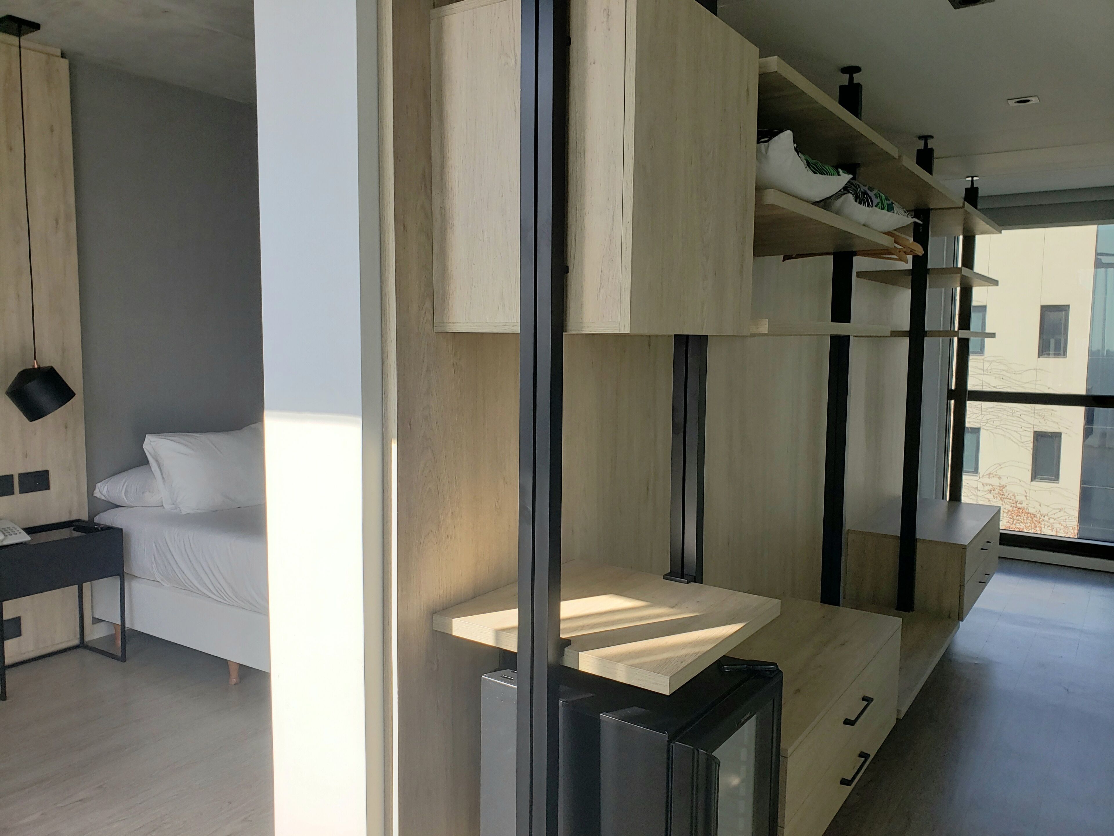 A modern hotel room with a minimalist design, featuring a bed, a desk, and built-in shelving with storage.