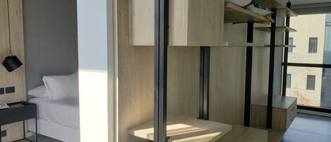 A modern hotel room with a minimalist design, featuring a bed, a desk, and built-in shelving with storage.