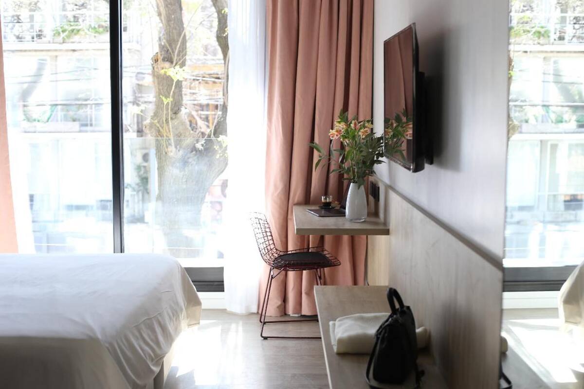 A hotel room with a bed, a desk with a chair, a TV mounted on the wall, and a view of the city through large windows.