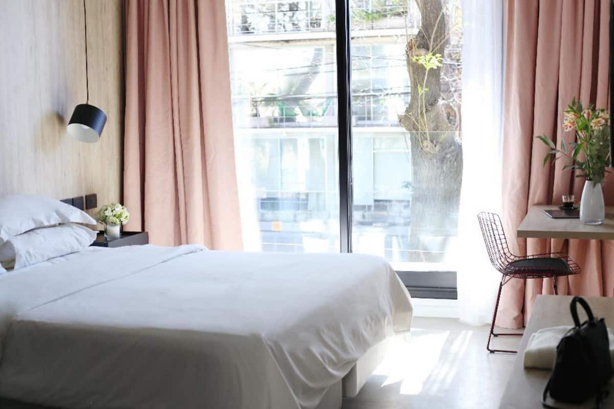 A modern hotel room with a large bed, a desk with a chair, a vase with flowers, and a view of buildings through the window.