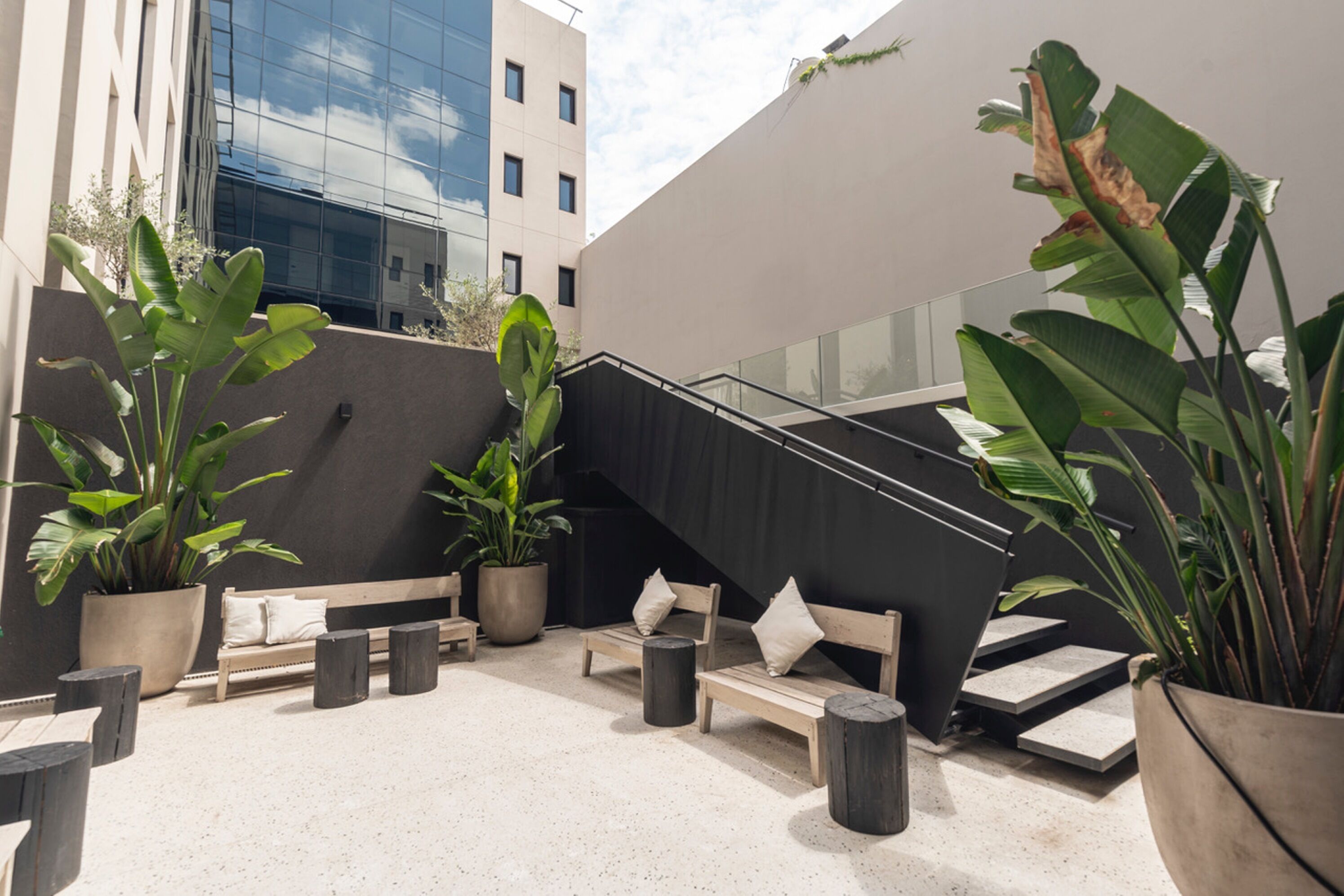 Un área de estar al aire libre con bancos y plantas en macetas, una escalera y un edificio moderno al fondo.