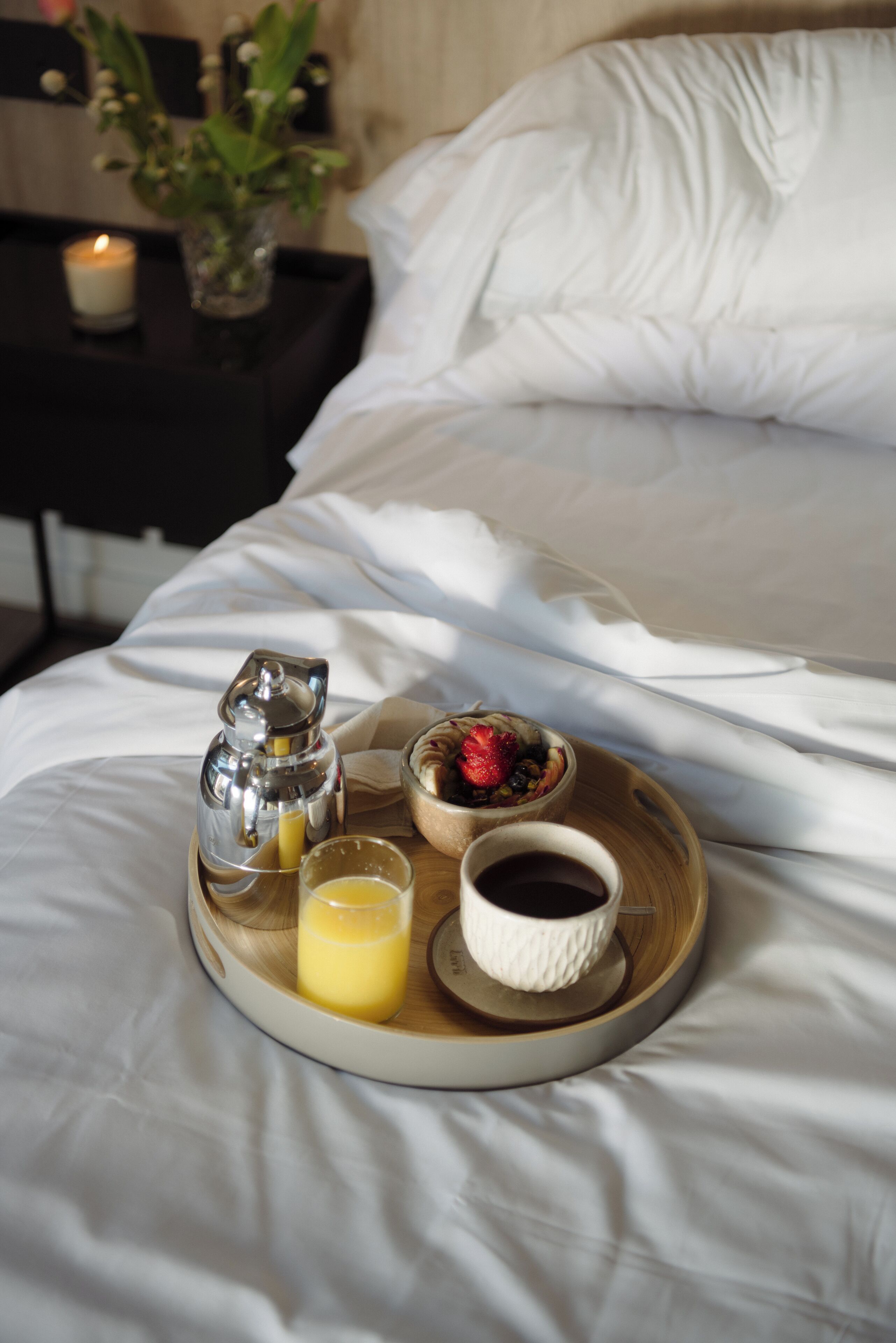 A breakfast tray with a coffee pot, a glass of orange juice, a bowl of fruit, and a cup of coffee on a bed.
