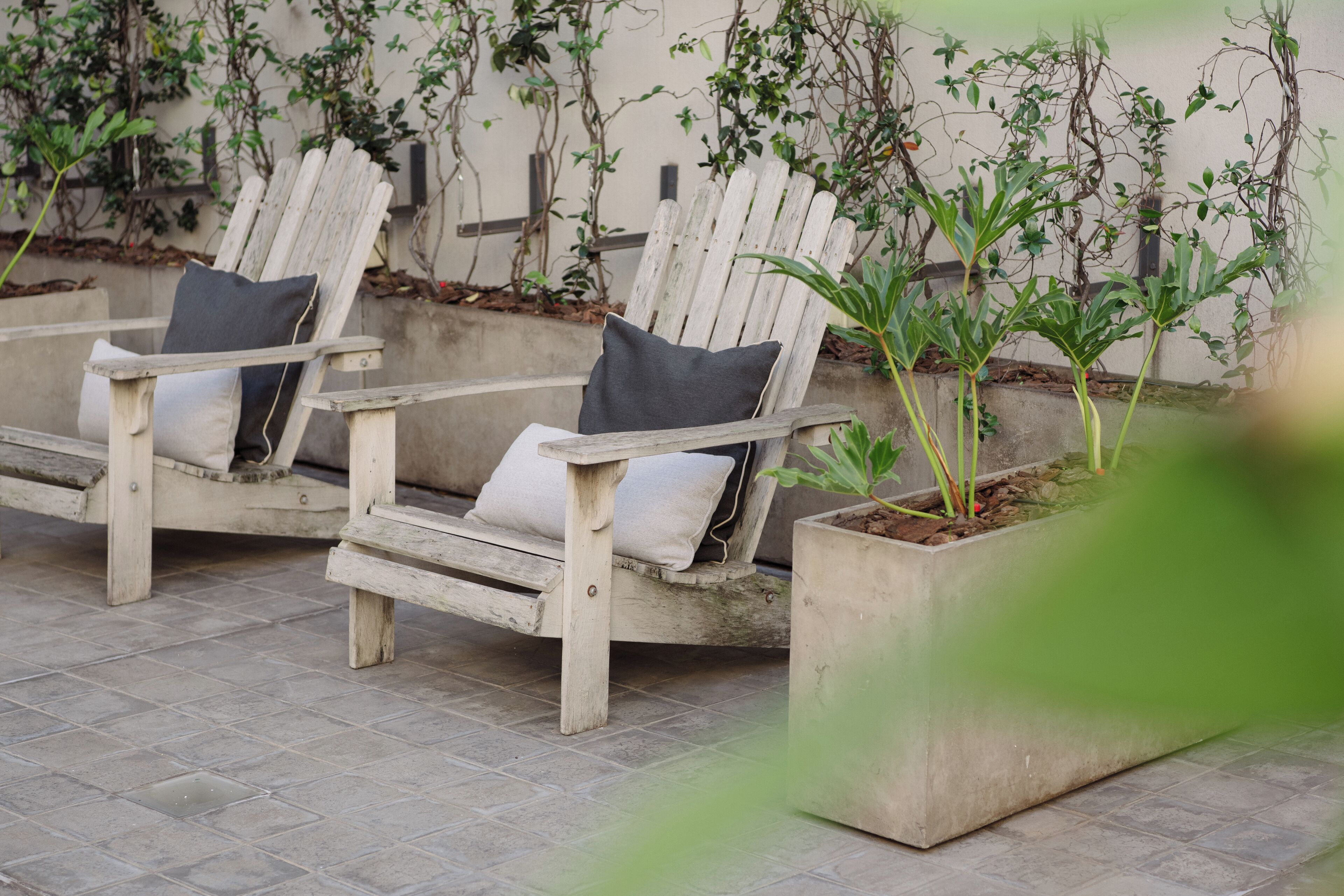Two wooden Adirondack chairs with cushions, positioned on a tiled patio with a concrete planter containing green plants.