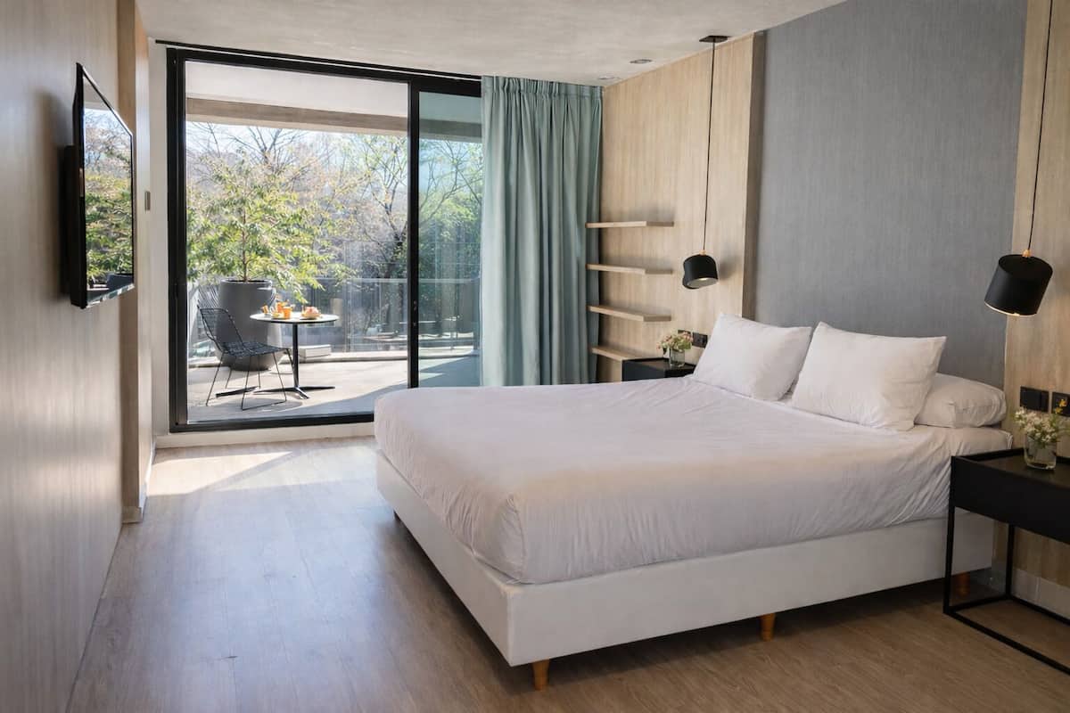 A modern bedroom with a large bed, a TV mounted on the wall, a bedside table with a lamp, and a view of an outdoor patio.
