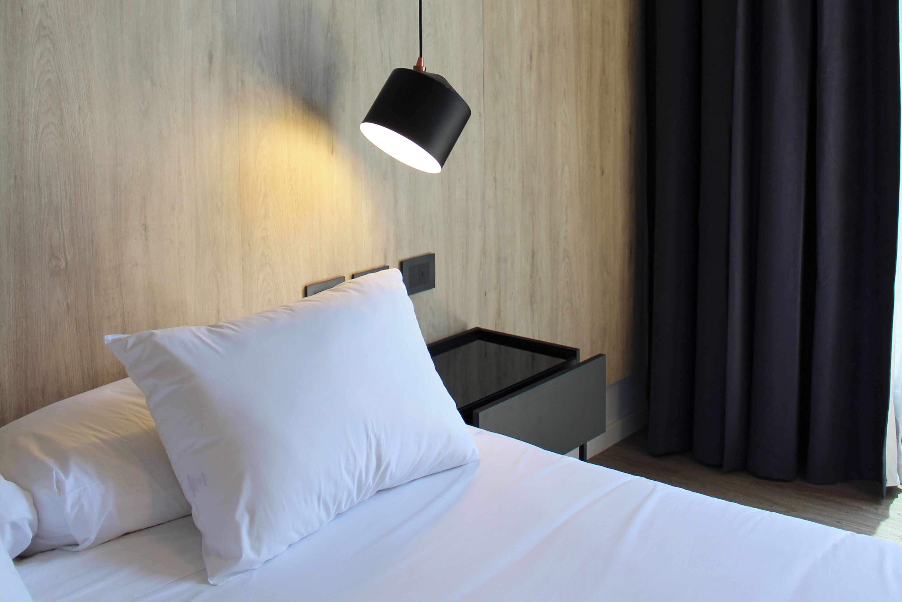 Una cama bien hecha con sábanas y almohadas blancas, una mesita de noche negra y una lámpara colgante.