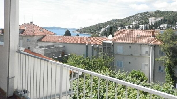 Un balcon offrant une vue sur une ville côtière, avec des maisons à étages aux toits rouges et une étendue d’eau visible au loin.