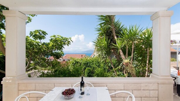 A terrace with a table set for two, a bottle of wine, and a bowl of fruit. The terrace overlooks a lush garden and a view of the sea. - Apartments Veve - Superior Studio Apartment with Terrace Sea View (Postira)