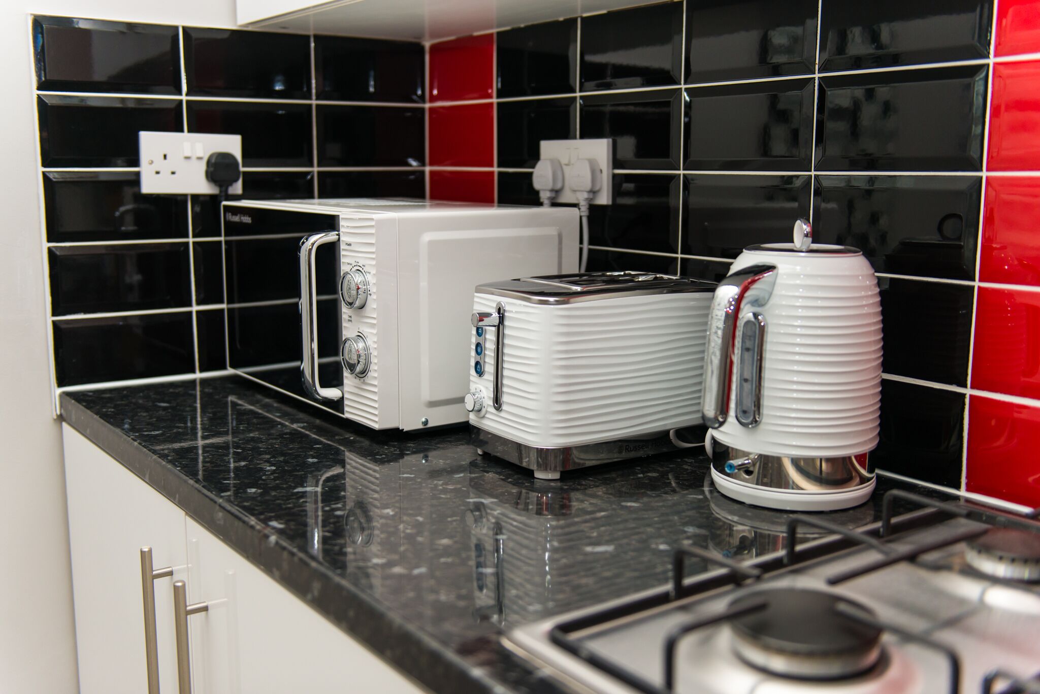 A kitchen with a black and red tiled backsplash, a white toaster, and a white electric kettle on a black countertop.