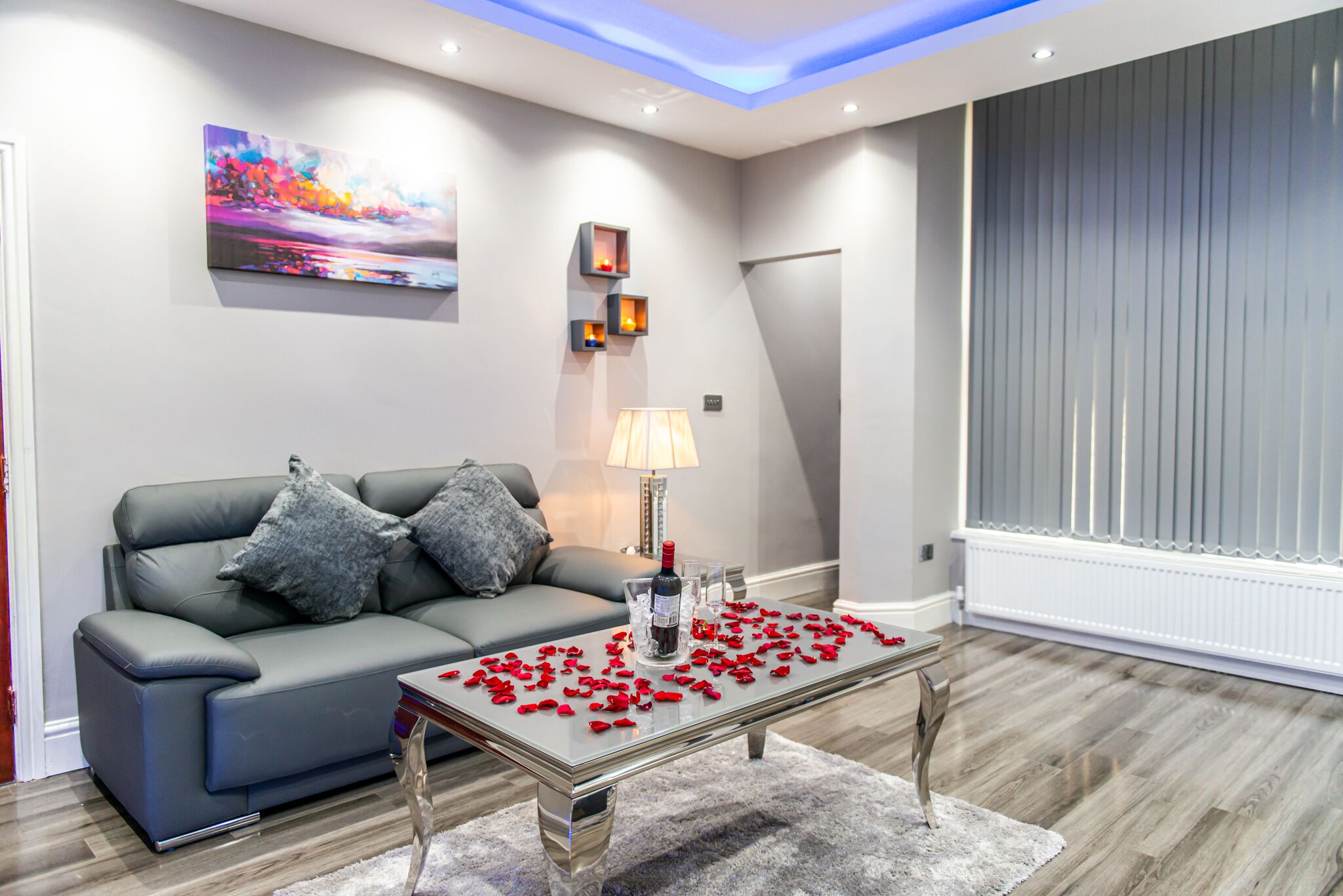 A modern living room with a grey sofa, a glass coffee table with red rose petals, a painting on the wall, and a window with blinds.