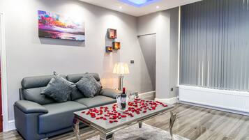 A modern living room with a grey sofa, a glass coffee table with red rose petals, a painting on the wall, and a window with blinds.
