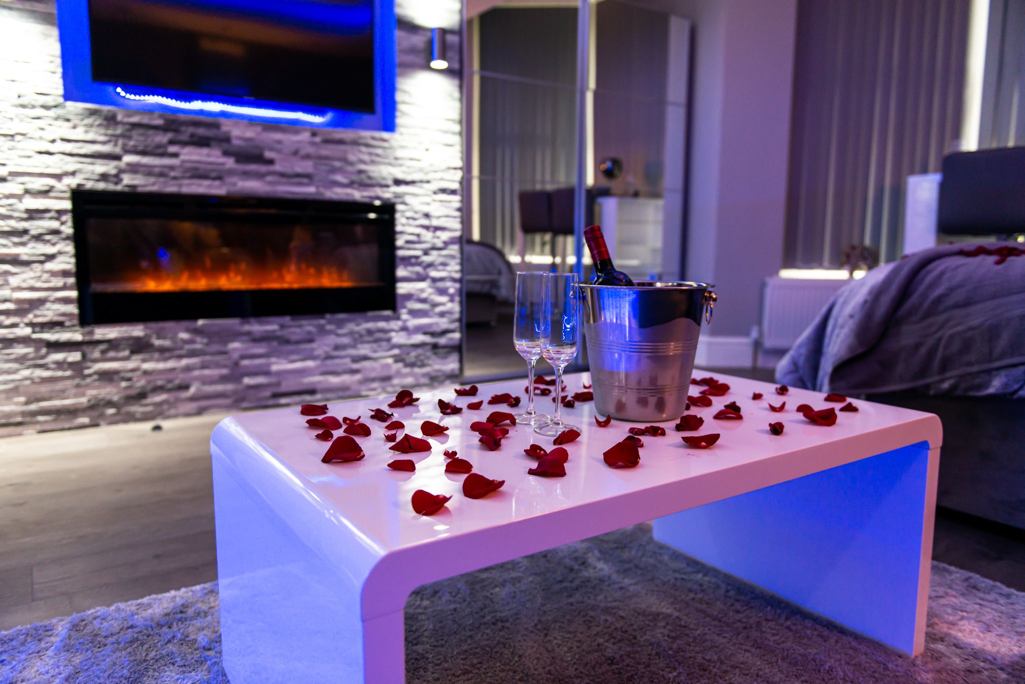 Ein modernes Wohnzimmer mit Steinwand, Kamin, Flachbildfernseher, weißem Tisch mit Rosenblüten und Eimer mit Gläsern.