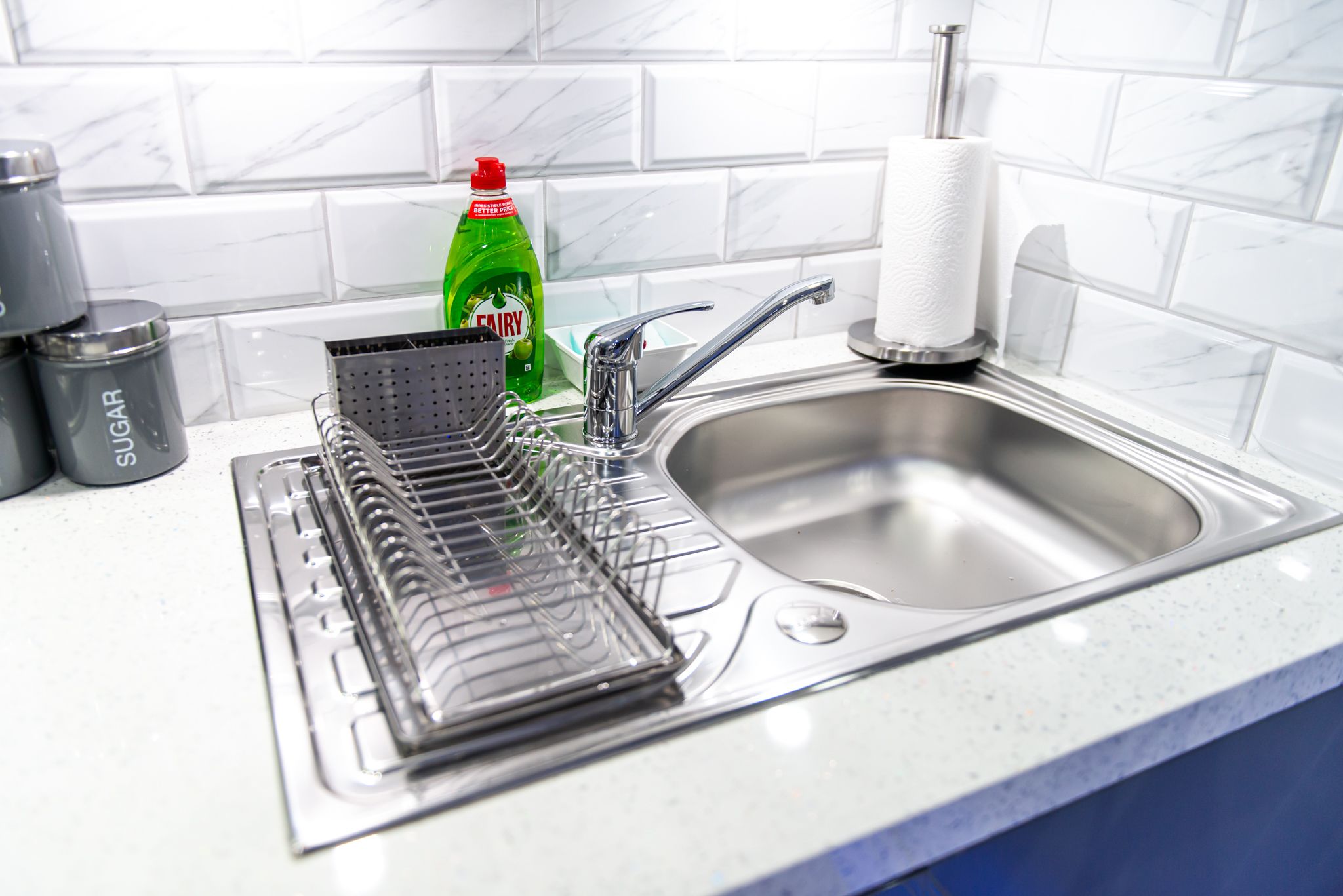 Una cocina con un escurreplatos, una botella de detergente Fairy y un recipiente para azúcar.