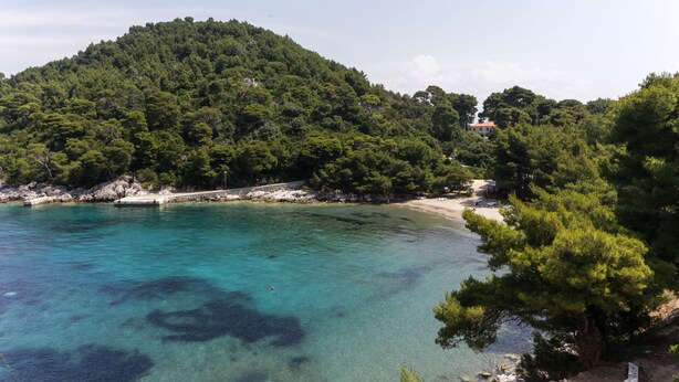 Eine Küstenlandschaft mit Strand, klarem türkisblauem Wasser und einem bewaldeten Hügel.