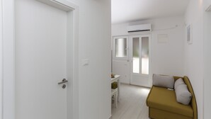 Ein gemütlicher Raum mit weißer Tür, einem kleinen Tisch mit Stuhl, einer Couch mit Kissen und einem Fenster mit satiniertem Glas.